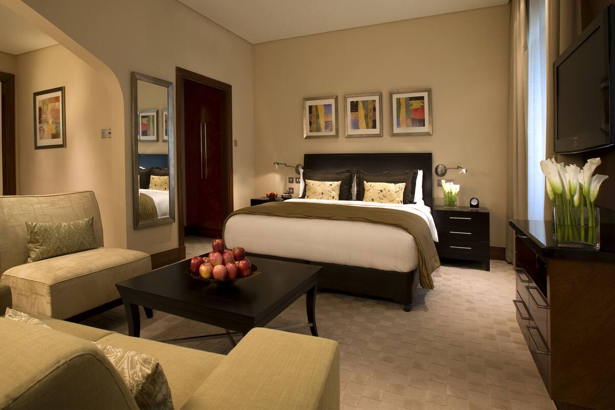 A hotel bedroom with a bed, a sofa, a coffee table with apples, a TV, and artwork on the walls.