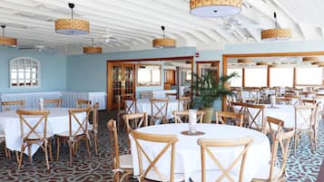 A spacious dining area with round tables and wooden chairs, featuring a patterned carpet and ceiling fans.