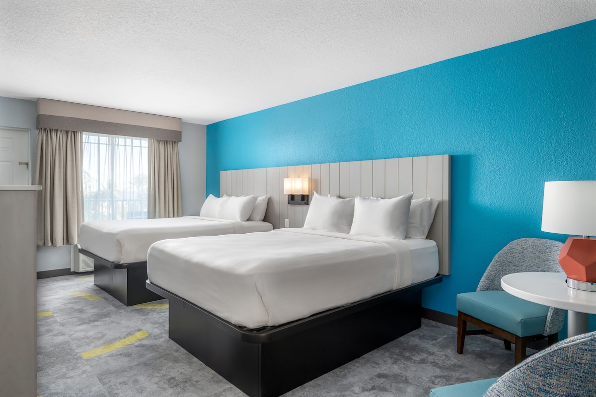 A hotel room with two beds, a blue accent wall, a round table with a lamp, and a view of the city through large windows.
