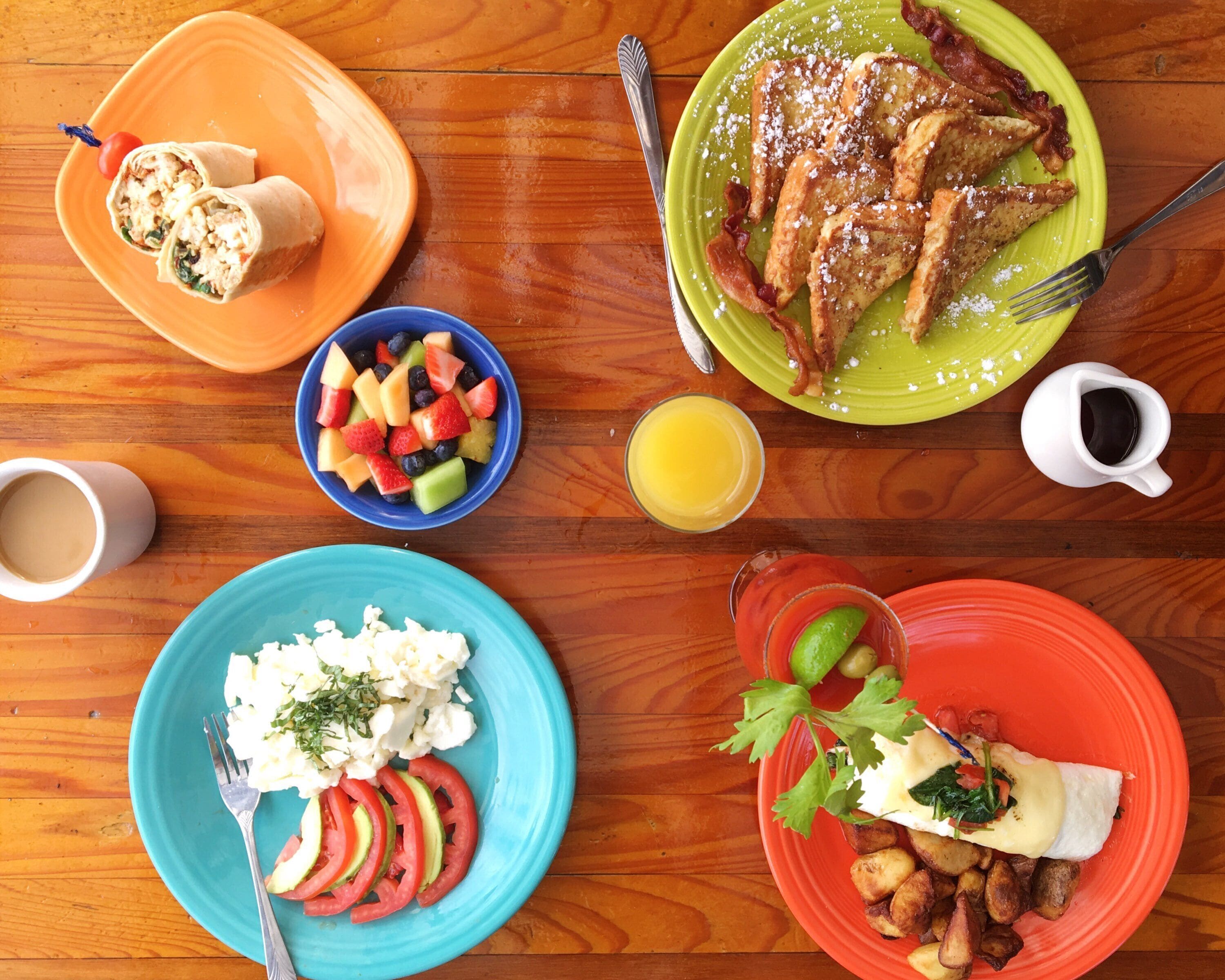 Una mesa de desayuno con varios platos, como panqueques, ensalada de frutas, huevos revueltos y papas asadas.