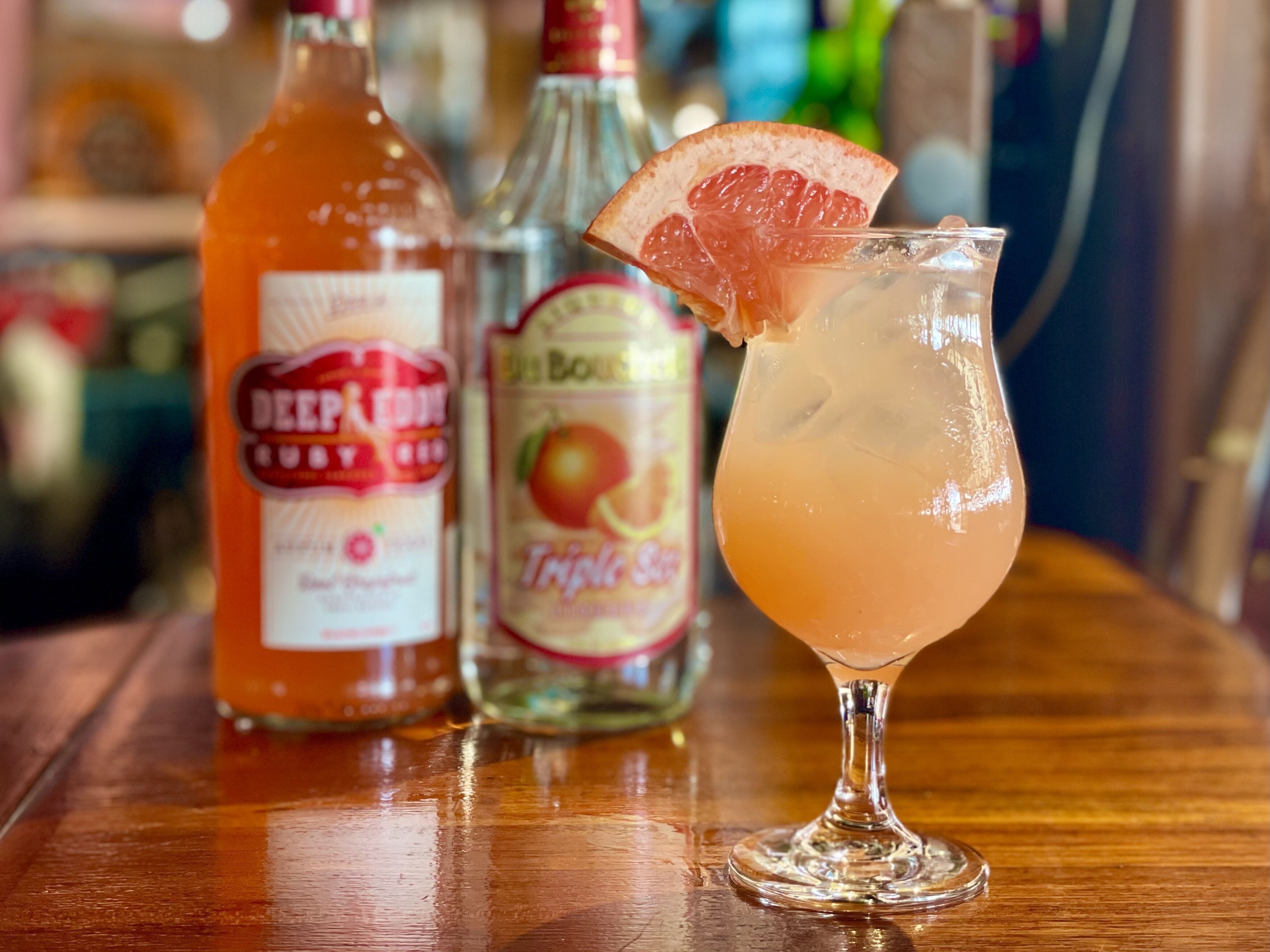 A cocktail glass with a citrus garnish on a wooden bar counter, accompanied by bottles of Deep Purple Gin and Triple Sec.