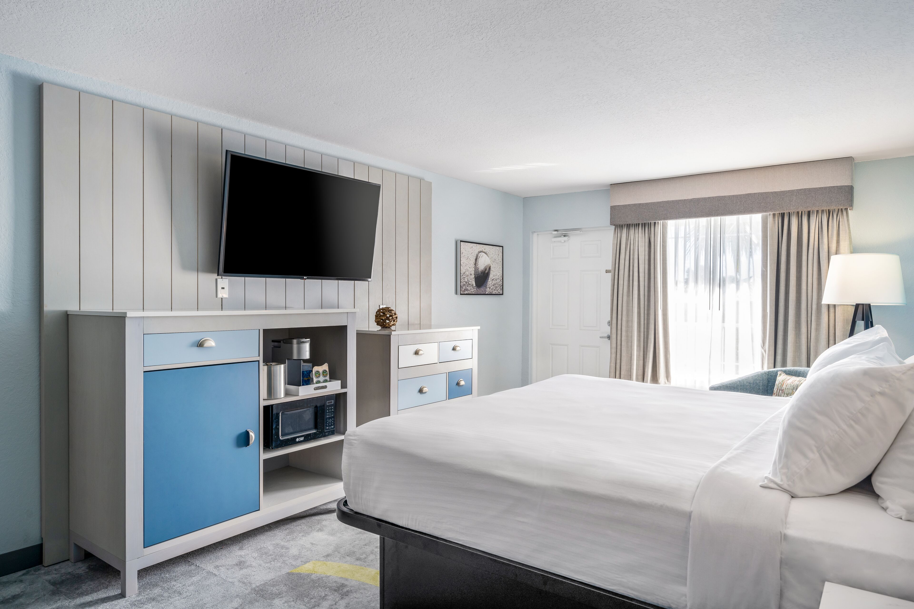Ein Hotelzimmer mit einem Bett, einem an der Wand befestigten Flachbildfernseher, einem Schreibtisch mit Lampe und einem fenster mit Vorhängen.