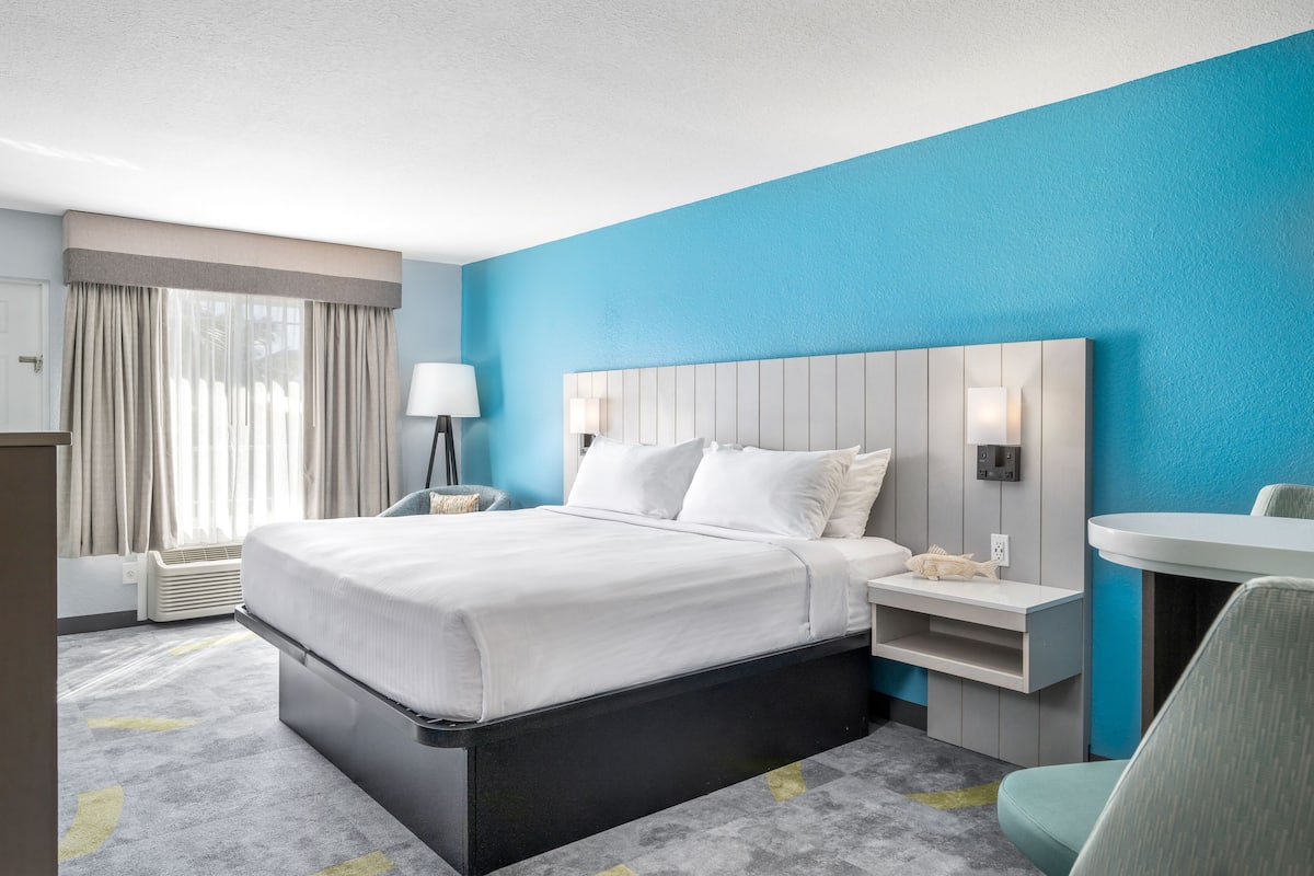 A hotel room with a bed, bedside table, lamp, and a small desk with a chair.