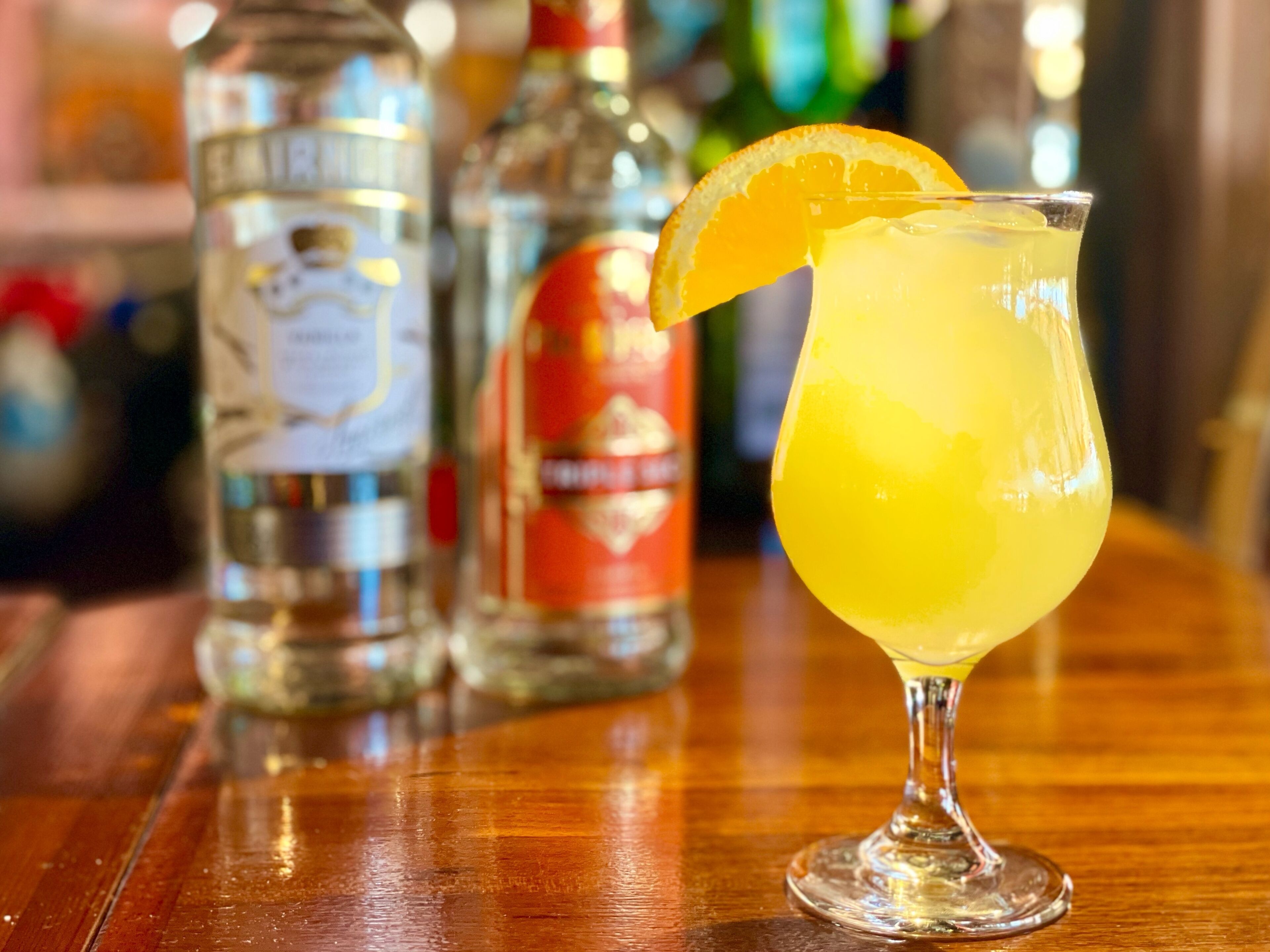 A cocktail glass with a lemon slice garnish on a bar counter, with bottles of liquor in the background.
