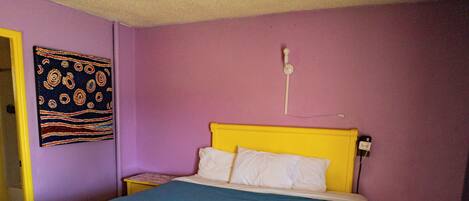 Cama con cubrecama azul, almohadas blancas y cabecero amarillo, además de un enchufe en la pared.