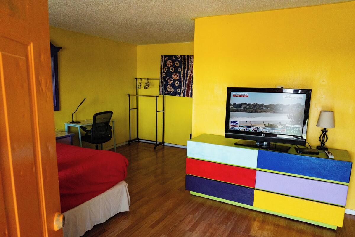 A hotel room with a bed, a desk with a chair, a TV on a colorful dresser, and a lamp.