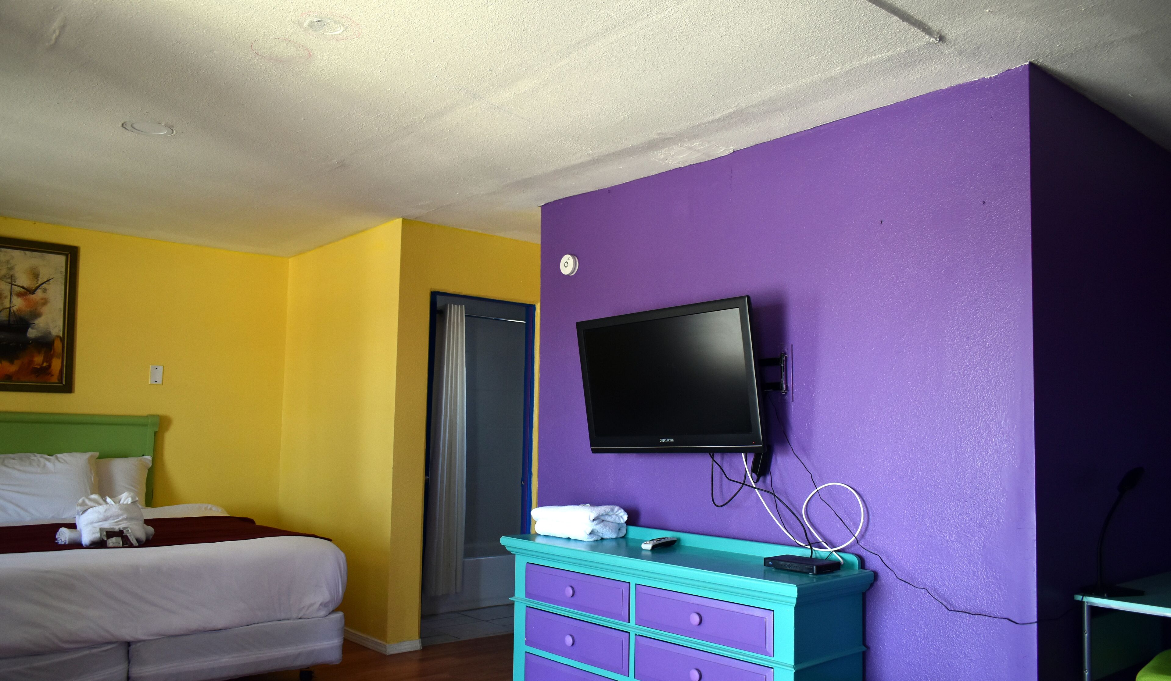 Une chambre d’hôtel avec un lit, un téléviseur fixé sur un mur violet et une commode turquoise.