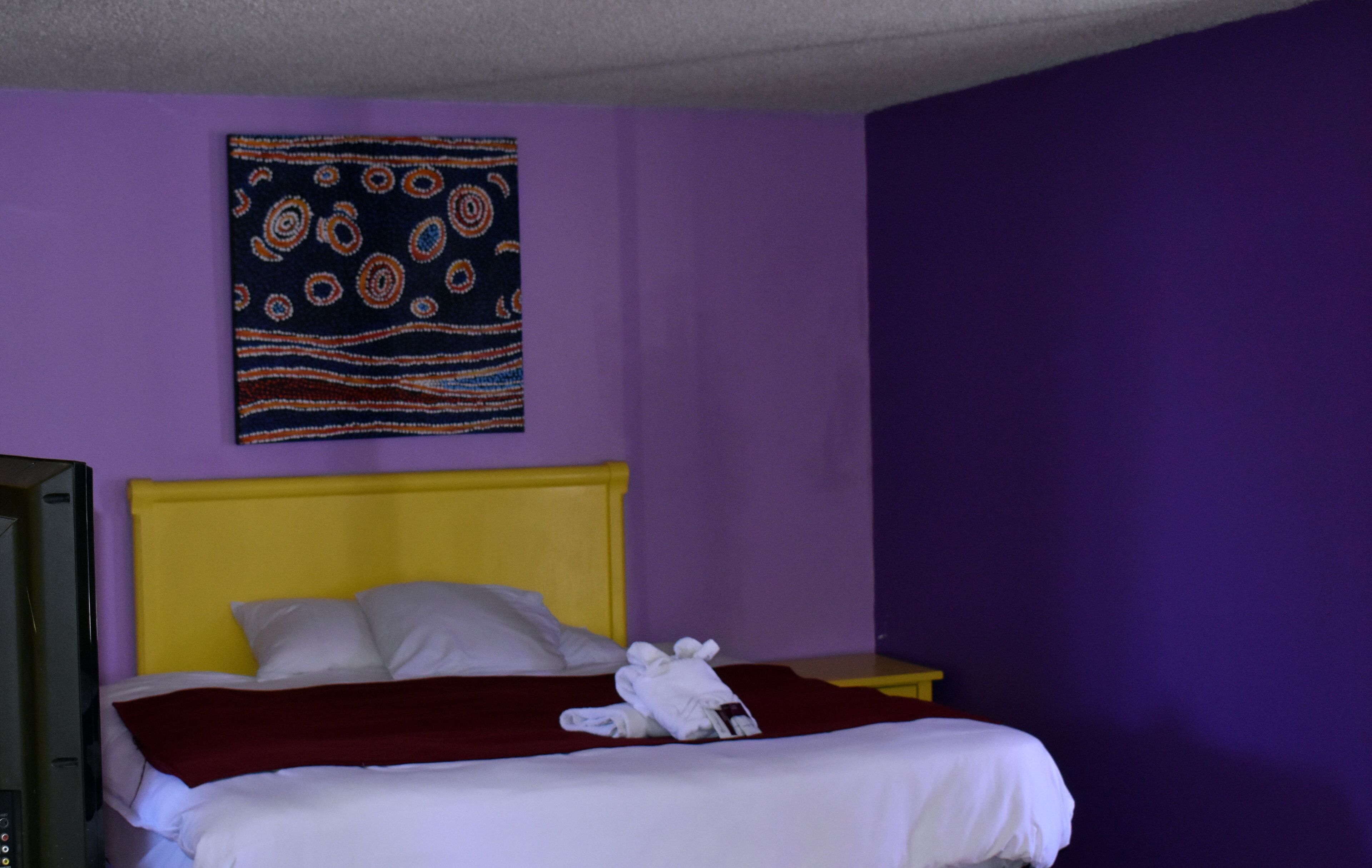 Une chambre d’hôtel avec un lit, une tête de lit jaune, un mur violet et une tapisserie décorative.
