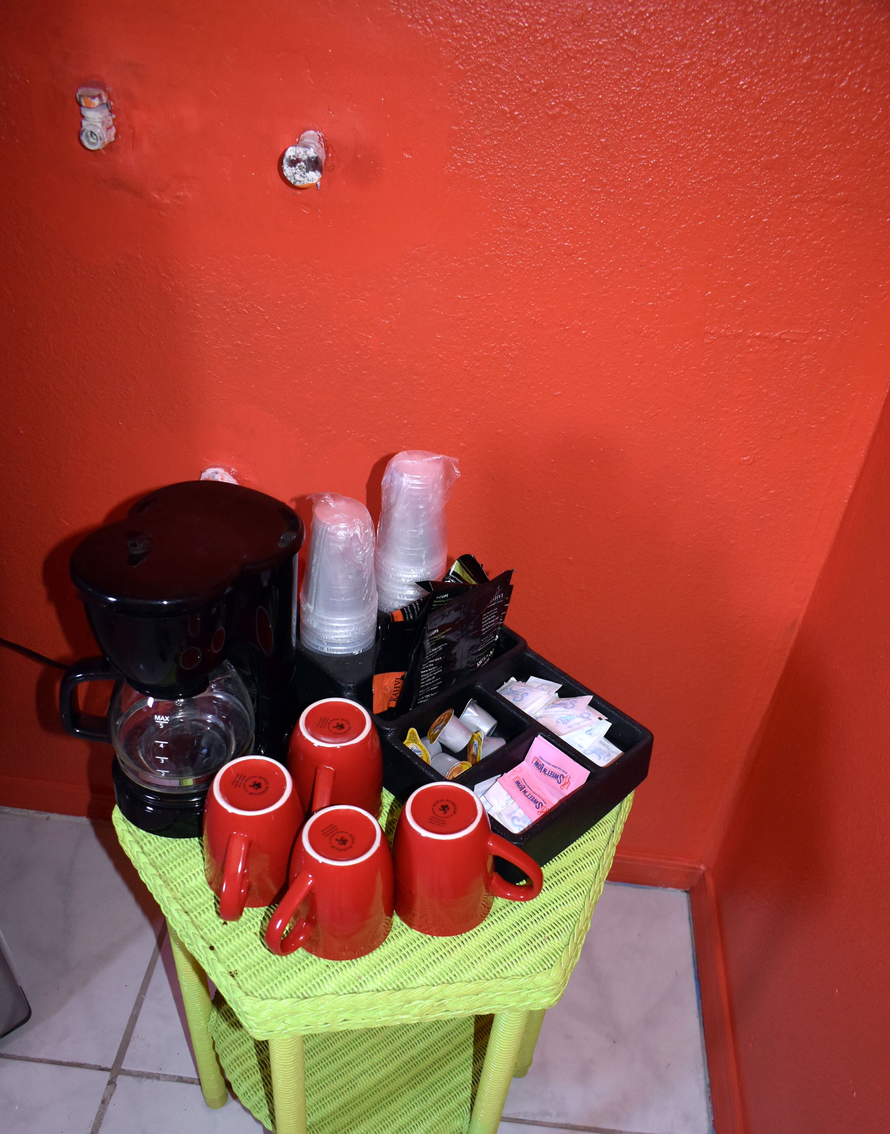 A small table with a coffee maker, red mugs, and a box of coffee pods against a red wall.