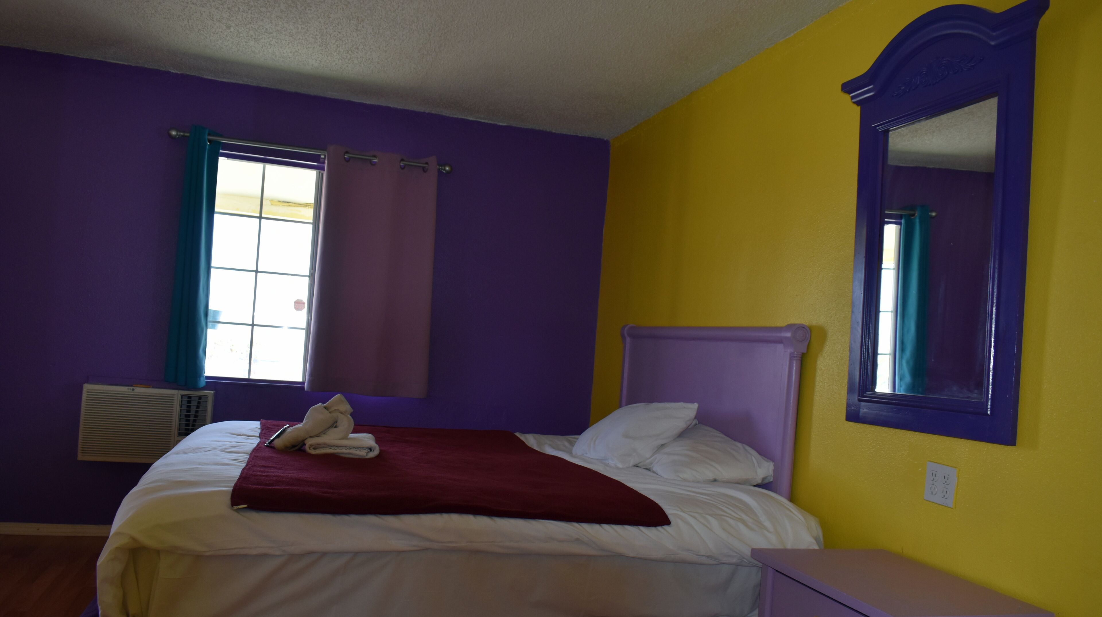 Un dormitorio con paredes moradas, una pared de acento amarilla, una cama con ropa blanca, una manta roja y una ventana con cortinas.