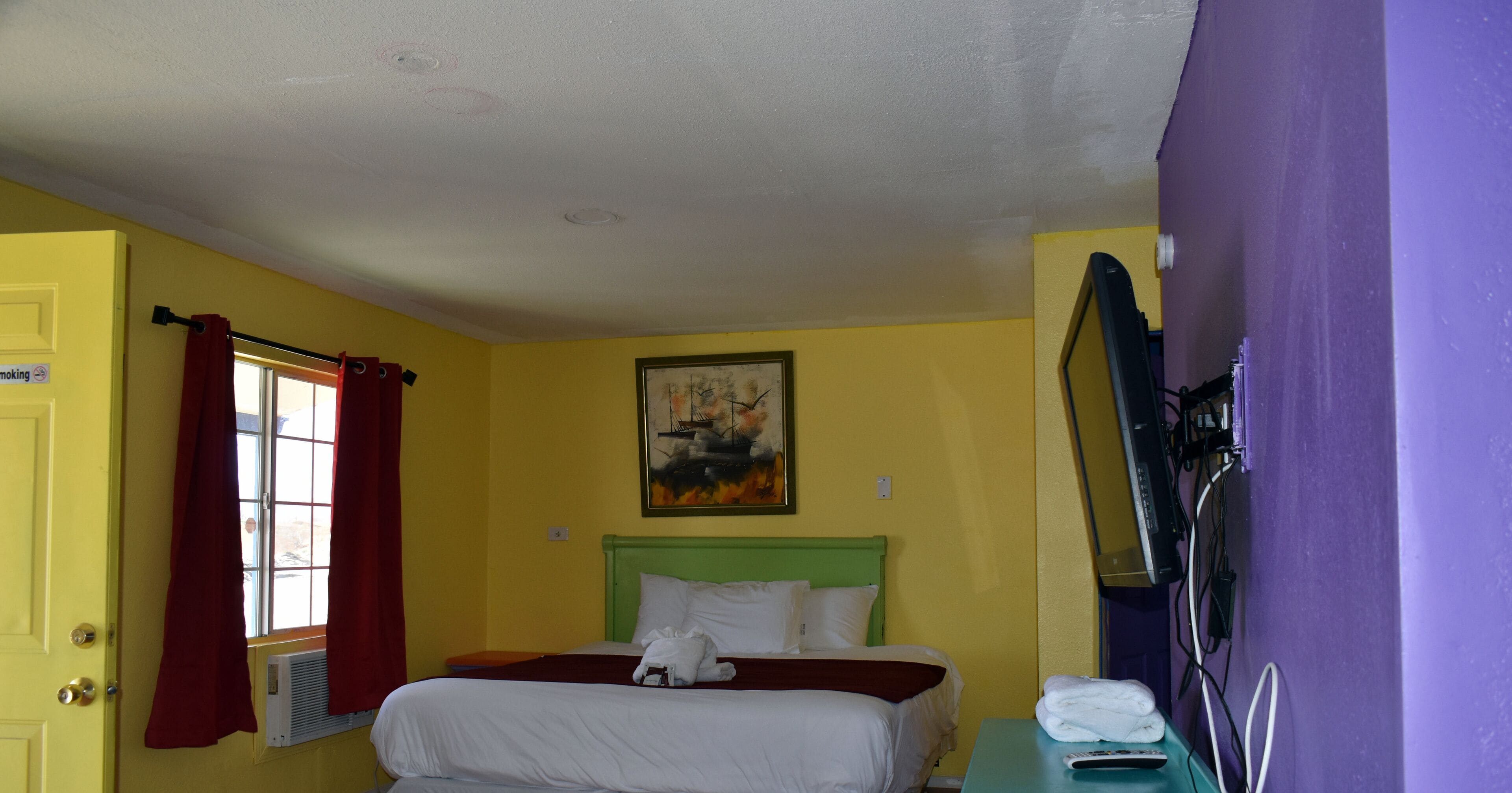 Une chambre d’hôtel avec un lit, un tableau au mur, une fenêtre avec des rideaux et un mur violet.
