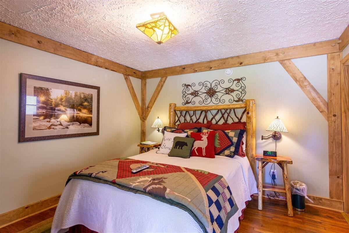 A bedroom with a wooden ceiling, a bed with a quilt, two bedside tables with lamps, a framed picture on the wall, and a clock on one of the tables.
