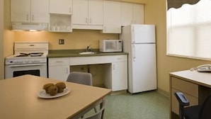 A compact kitchen with white cabinets, a microwave, a refrigerator, and a table with a plate of food. - Glendenning Hall (Charlottetown)
