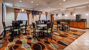 A hotel dining area with a large table, chairs, a patterned carpet, a kitchen area with cabinets and appliances, and a television mounted on the wall. - Best Western Jacksonville Inn (Jacksonville)