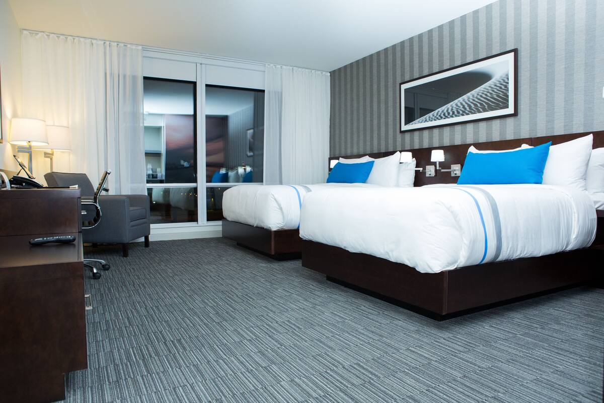 Ein Hotelzimmer mit zwei Betten, einem Schreibtisch, einem Stuhl und einem großen Fenster mit Vorhängen.