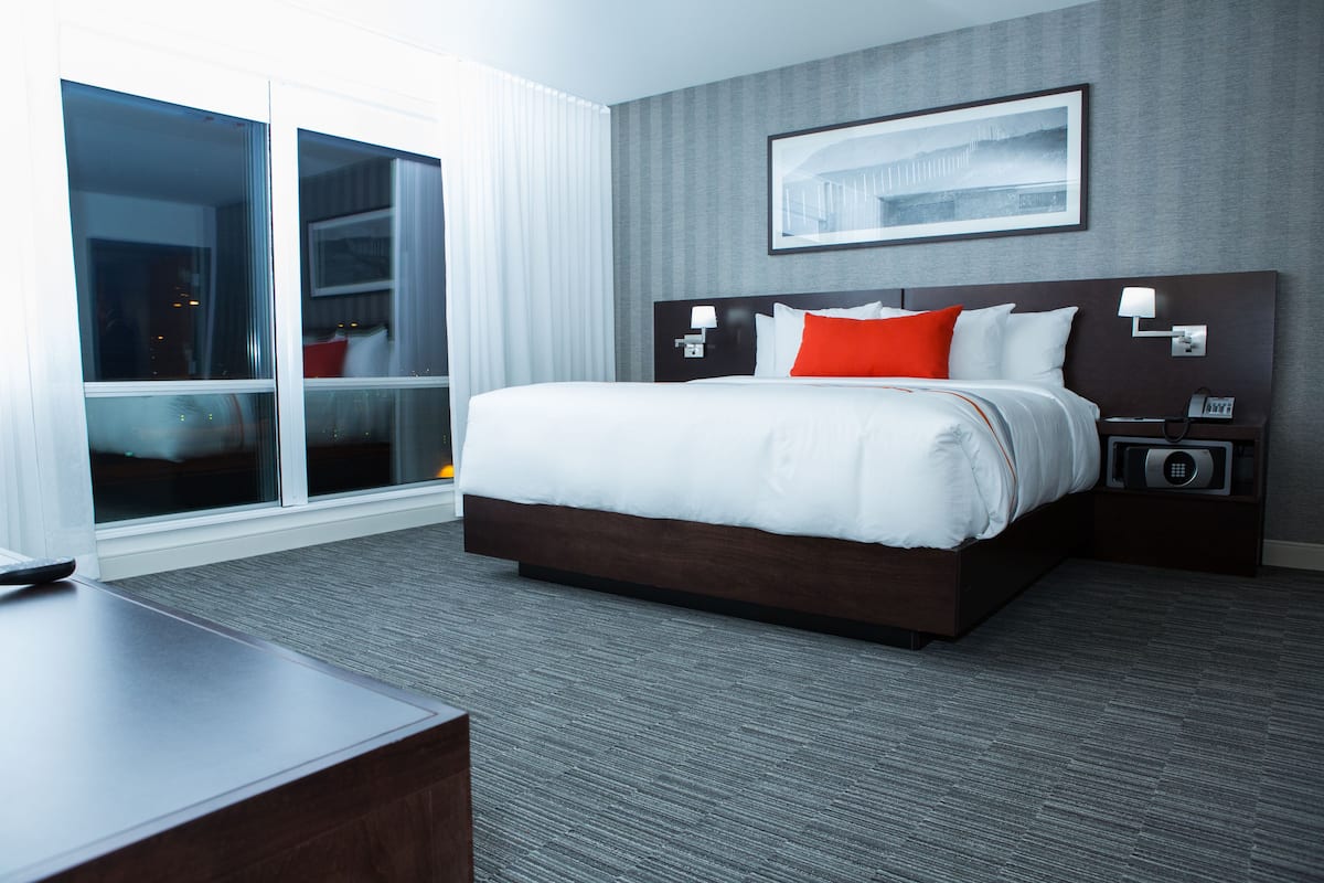 Ein Hotelzimmer mit einem großen Bett, einem Schreibtisch, einem Nachttisch, einem Telefon und einem Fenster mit Jalousien.