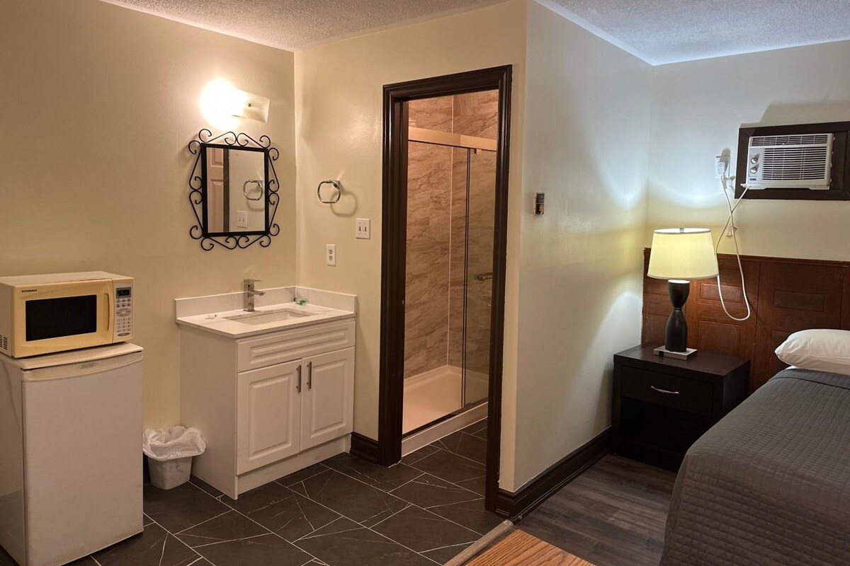 Una habitación de hotel con baño, una cama, una mesita de noche, una lámpara y un microondas.