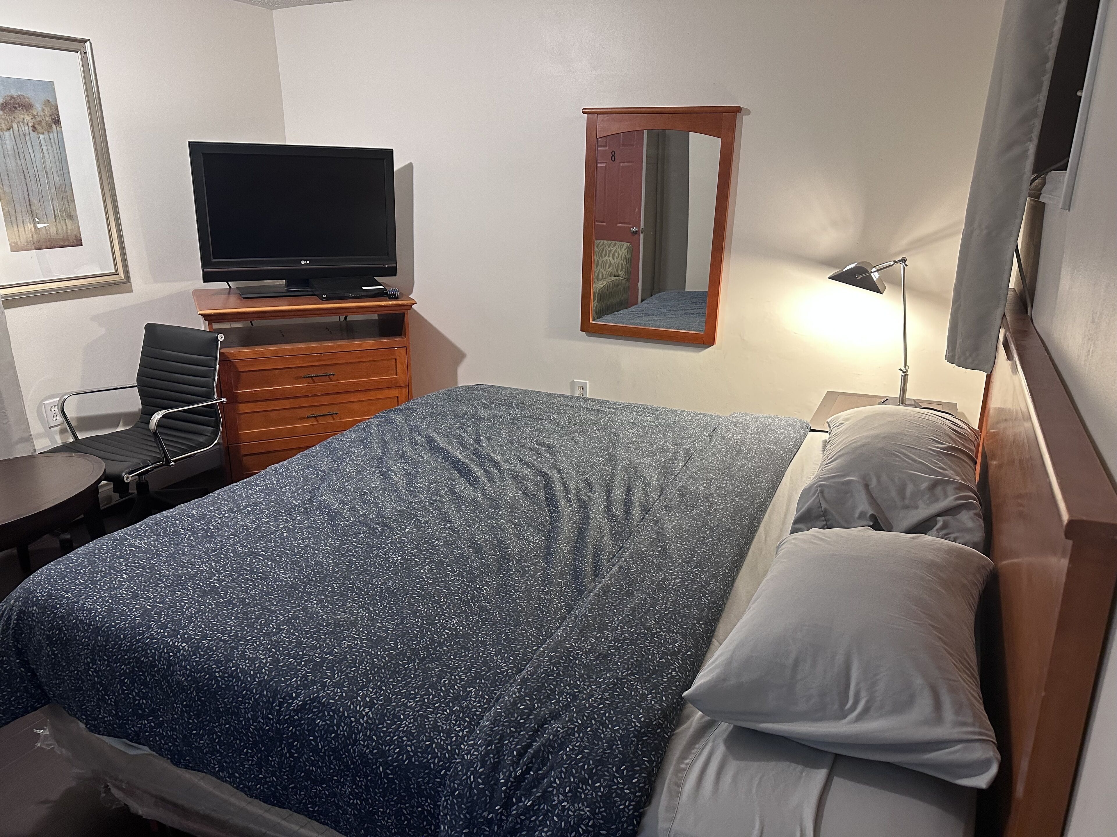 Un dormitorio con cama, escritorio y silla, televisión, cómoda y espejo.
