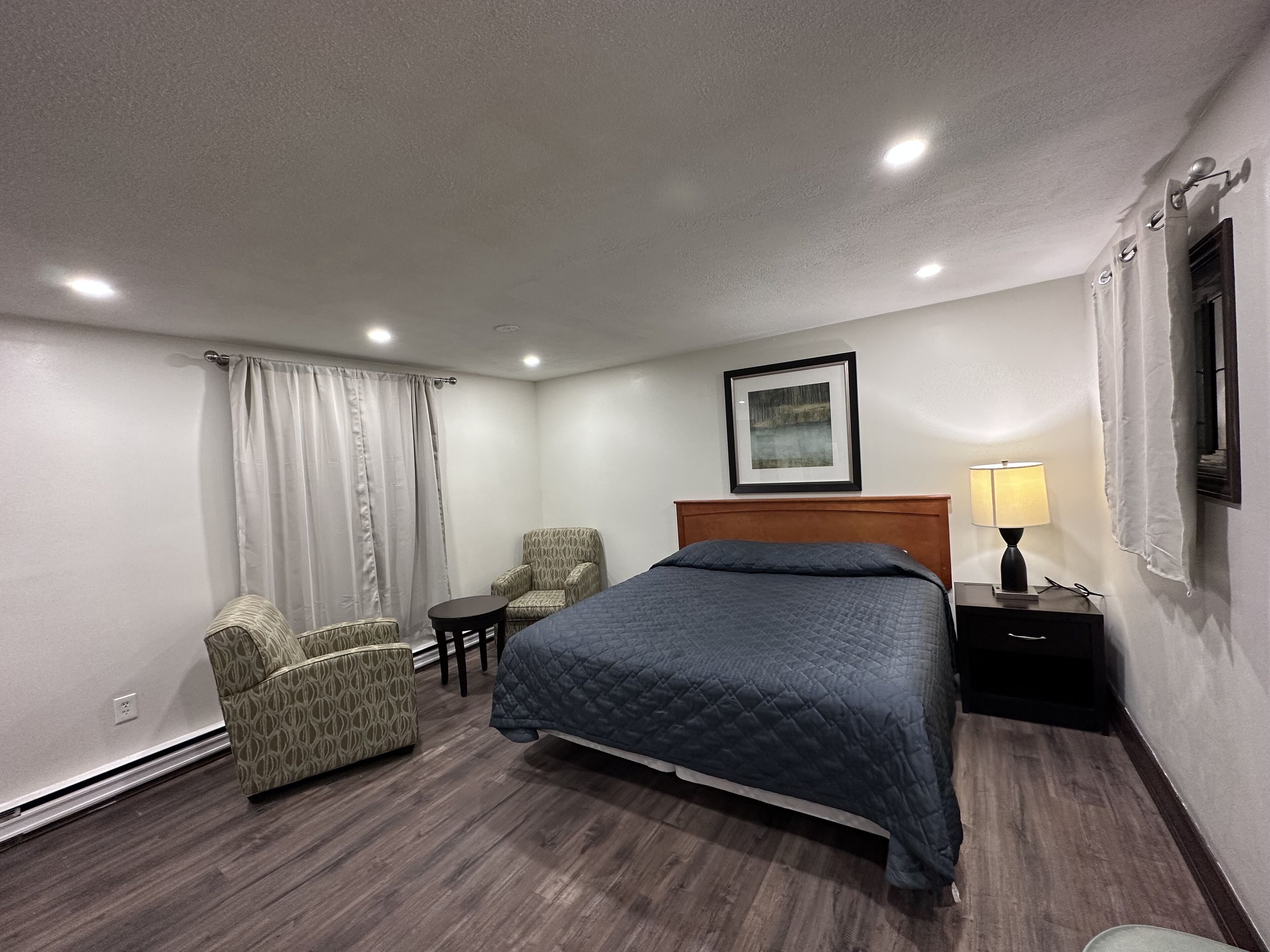 Habitación de hotel con cama, una silla, una mesita, una lámpara y un cuadro en la pared.