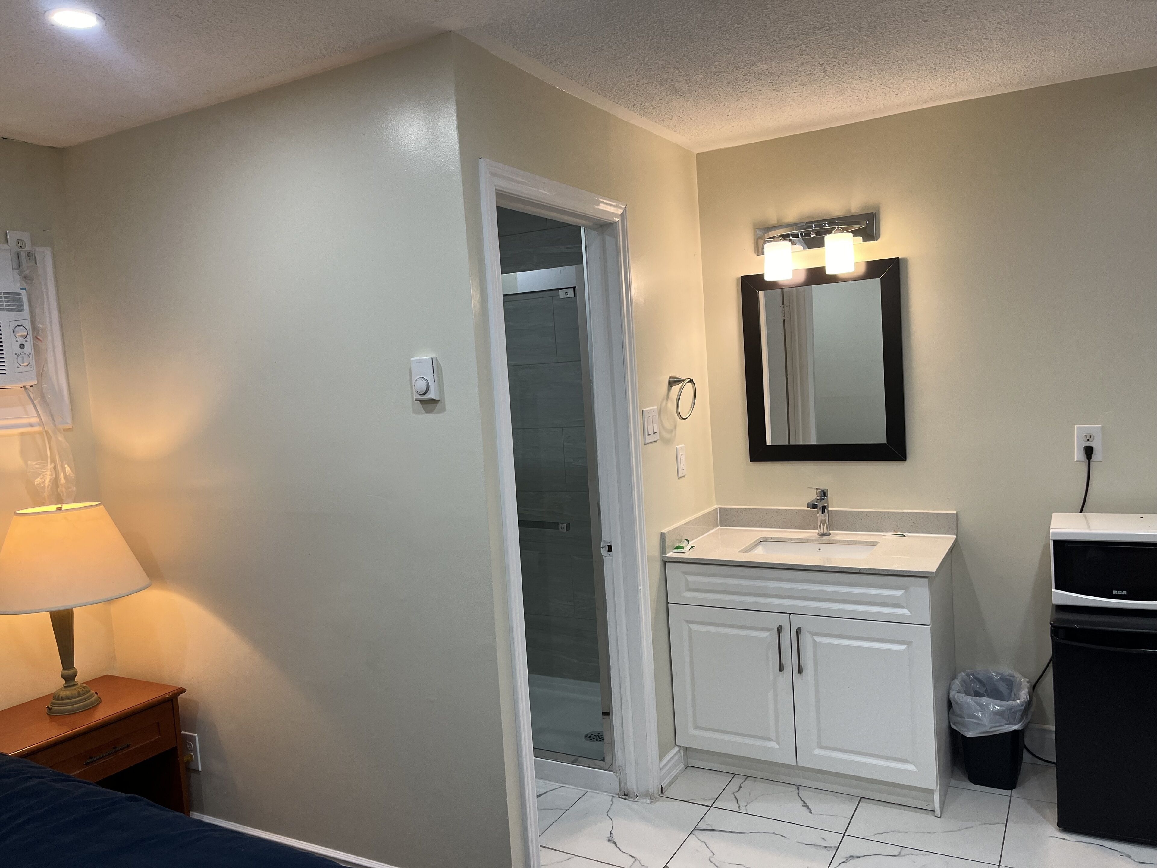 Un baño con lavamanos, espejo y mueble blanco, puerta de ducha y un cubo de basura.