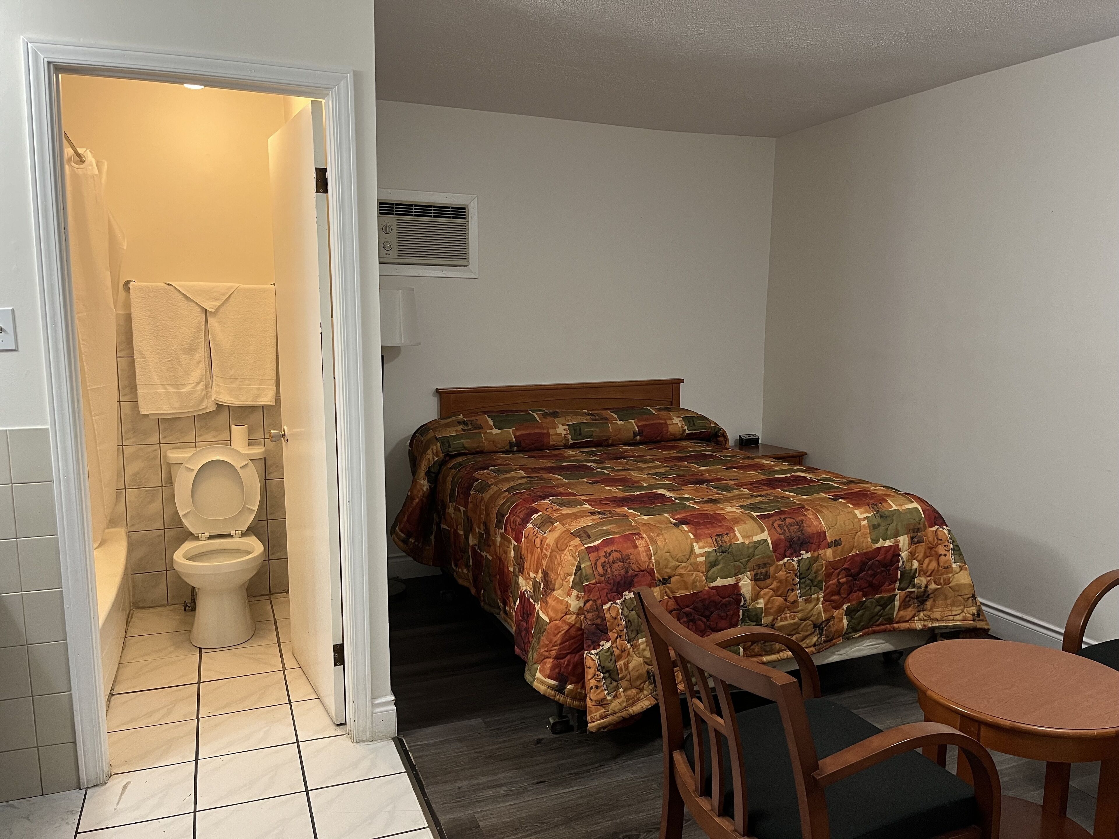 A bedroom with a bed, a wooden chair, a small table, a bedside lamp, and a bathroom with a toilet and towel rack.
