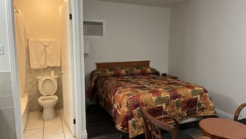 Un dormitorio con cama, una silla de madera, una mesita pequeña, una lámpara de noche y un baño con inodoro y perchero para toallas.
