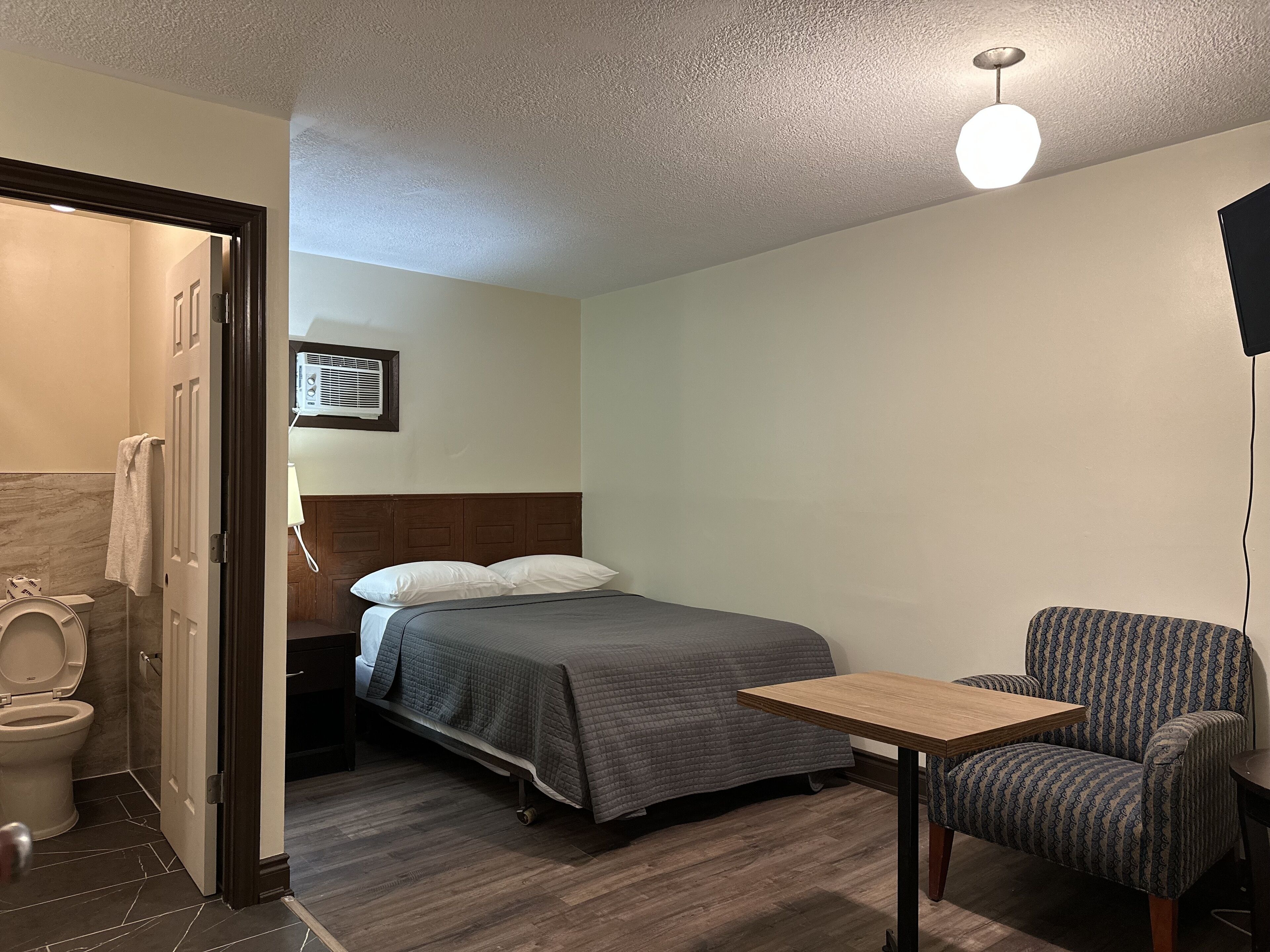 A hotel room with a bed, a chair, a table, a toilet, and a ceiling light.