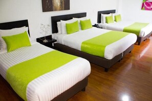 Hotellihuone, jossa on kolme sänkyä. Jokaisessa sängyssä on valkoinen ja vihreä peitto, ja huoneen lattia on puuta.
