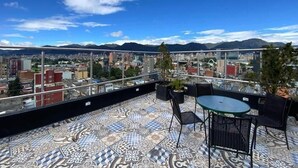 A rooftop terrace with patterned flooring, a table, chairs, and a view of a cityscape with mountains in the background. - Hotel Macao Colombia (Bogotá)