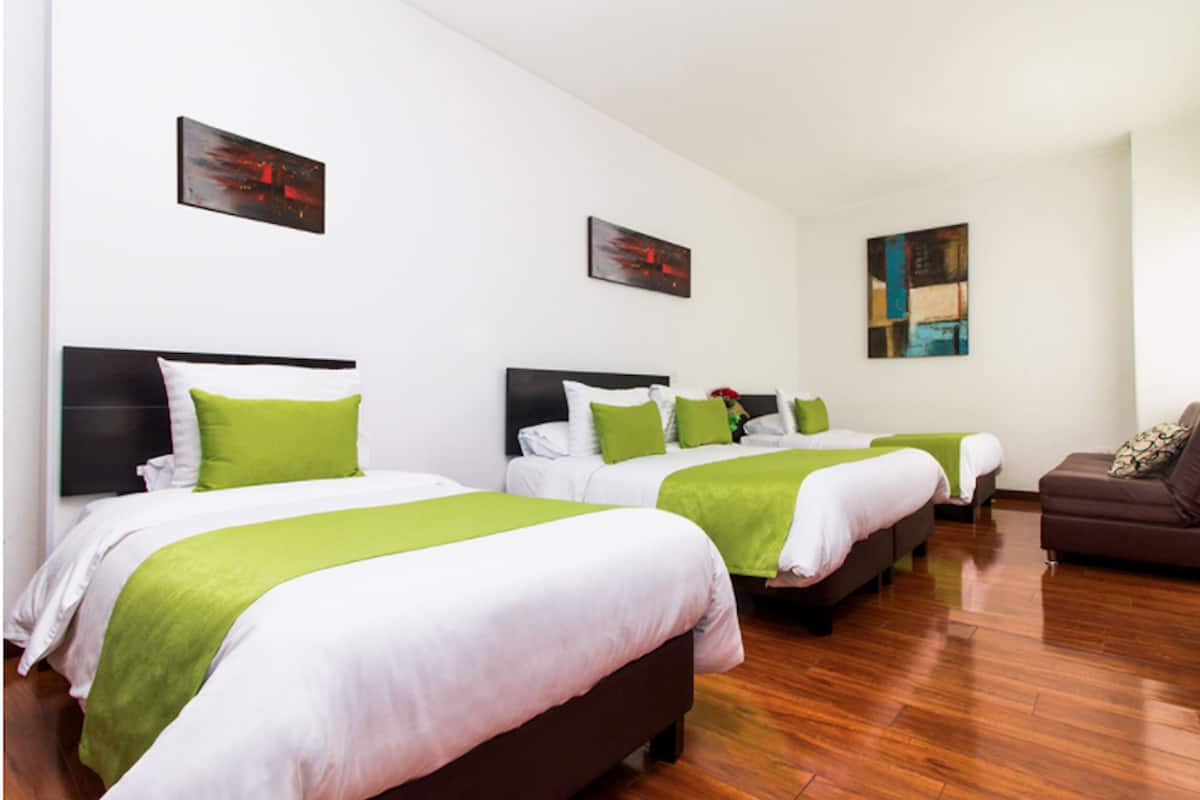 Habitación de hotel con tres camas, cada una con ropa de cama blanca y detalles verdes, un sofá marrón y obras de arte en las paredes.