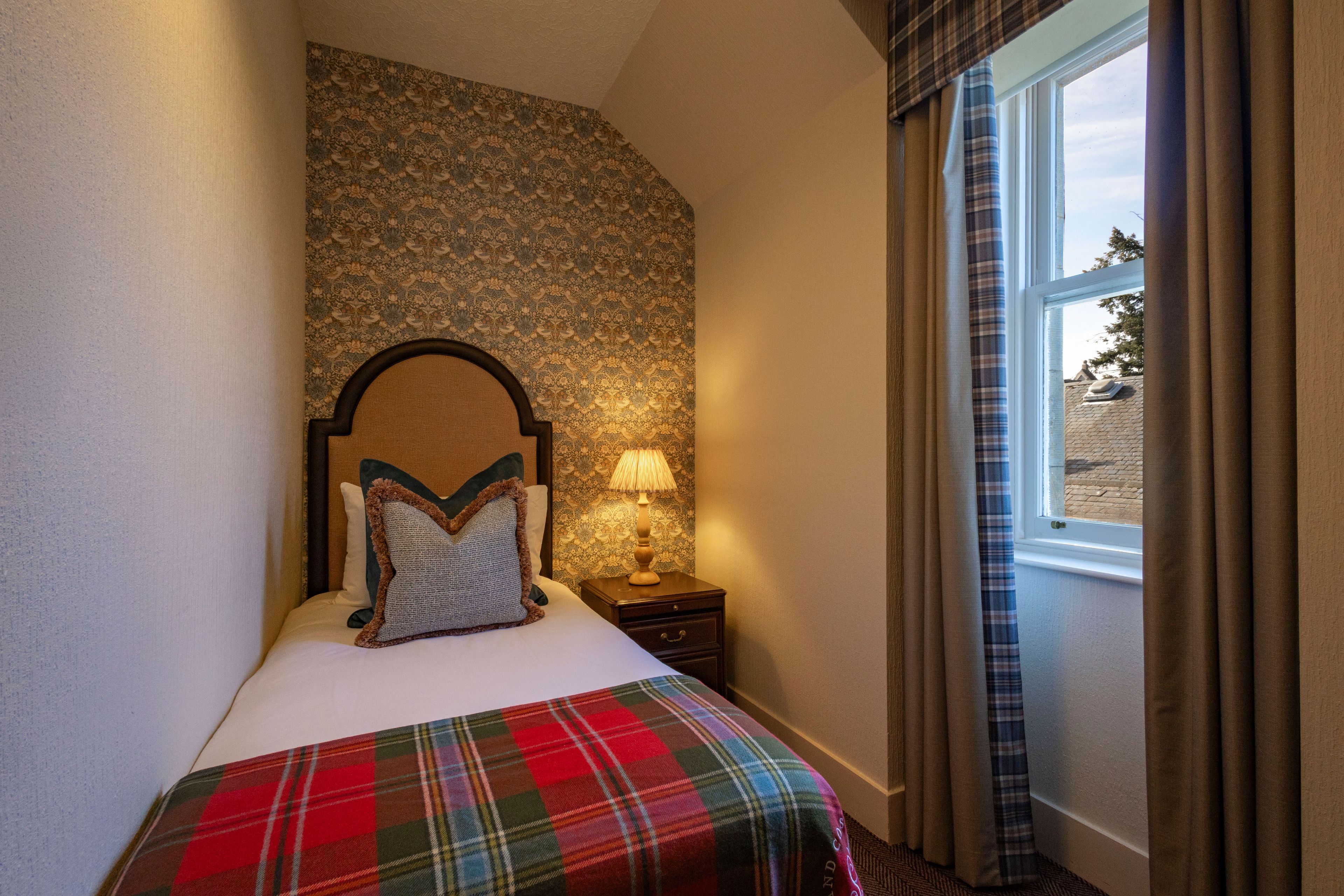 Una habitación de hotel con cama, mesita de noche, lámpara y ventana con cortinas.