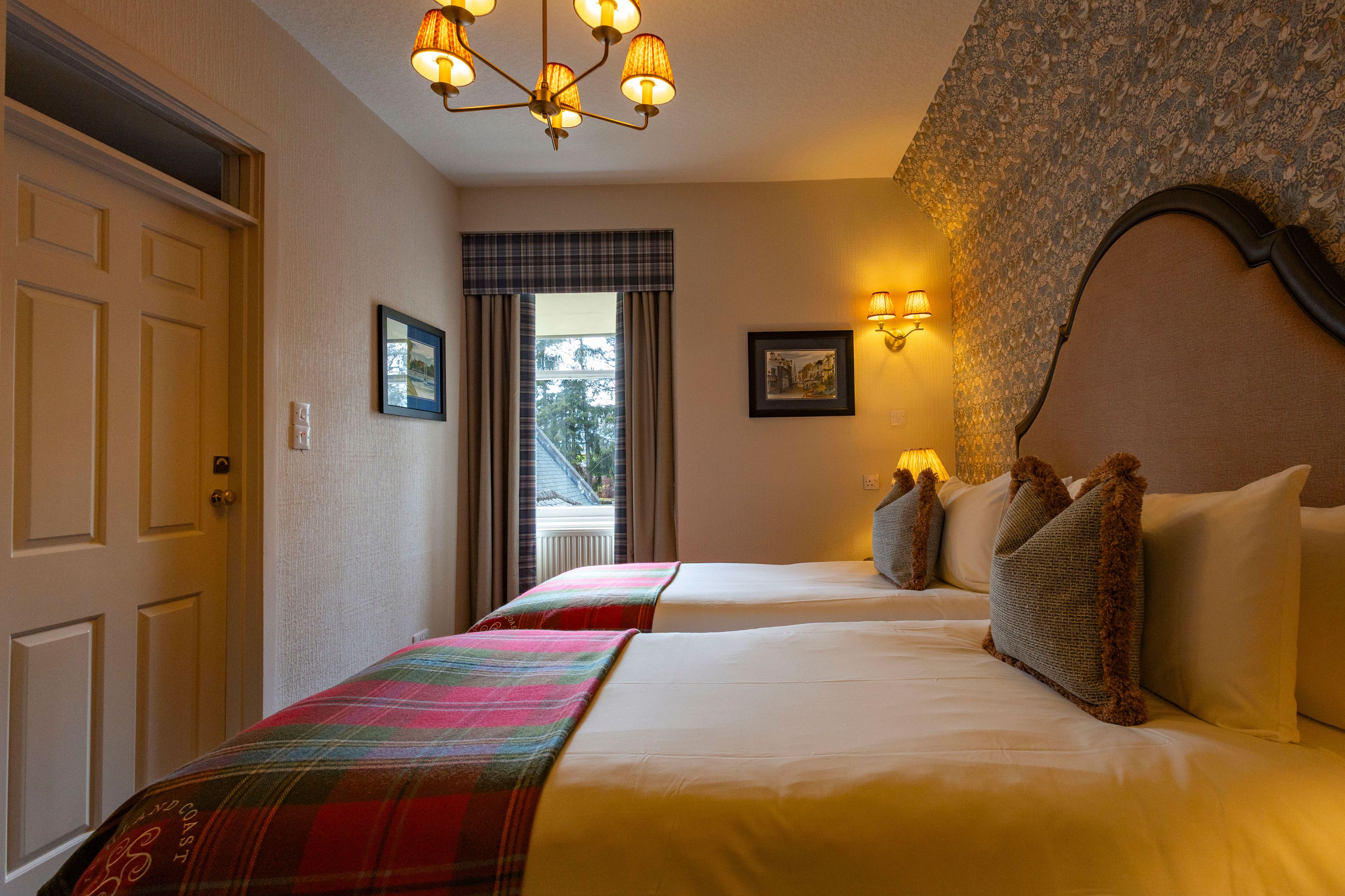 A hotel room with two beds, a chandelier, a window with curtains, and a wall-mounted lamp.