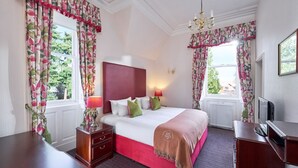 A hotel room with a large bed, two bedside tables, a desk, and a chair. There are two windows with curtains, a chandelier, and a radiator. - Lochardil House Hotel (Inverness)