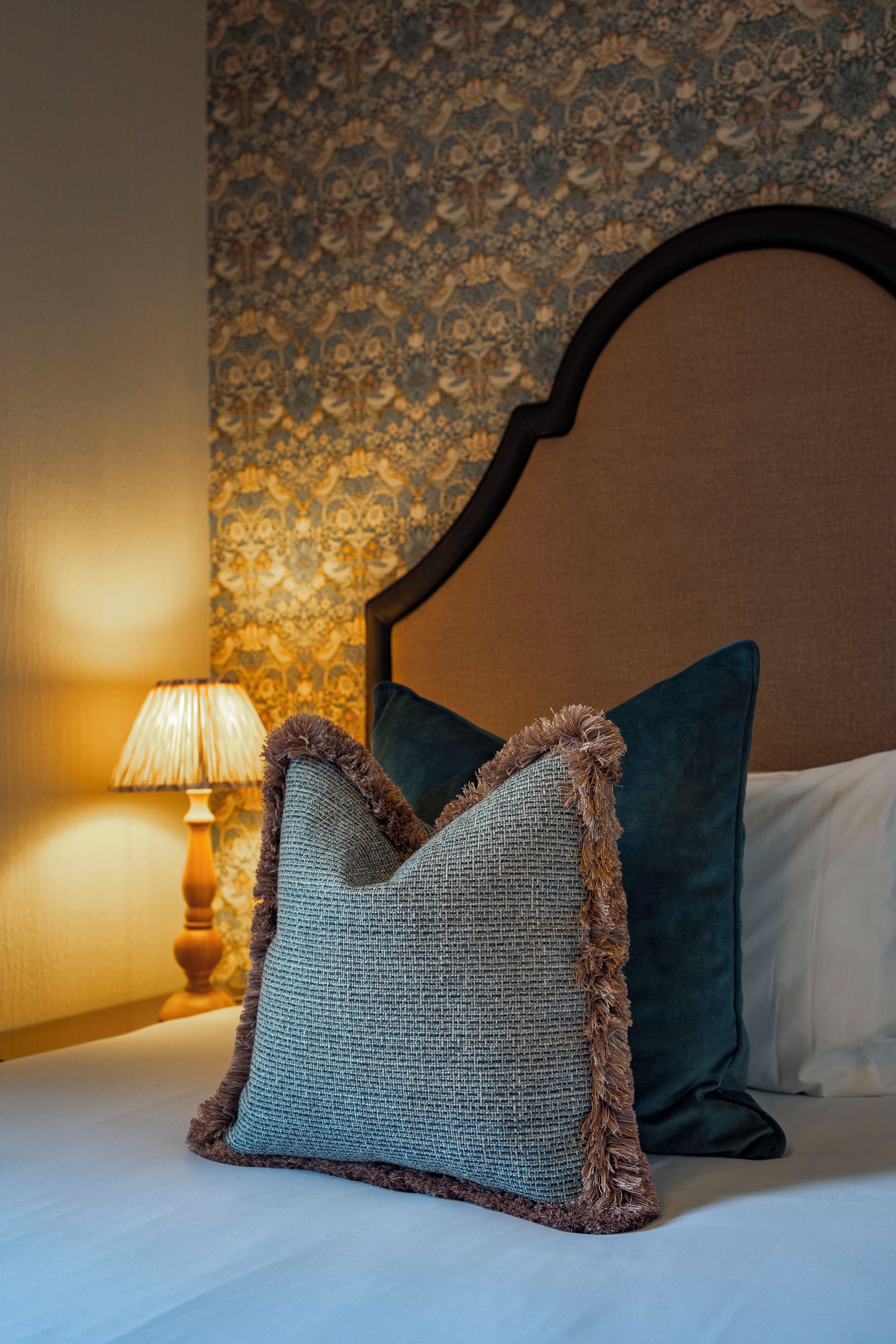 Una cama con dos almohadas, una lámpara y un papel tapiz estampado.