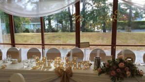 A wedding reception table with 'Mr & Mrs' illuminated letters, a floral centerpiece, and a view of a grassy area through large windows. - Lochardil House Hotel (Inverness)