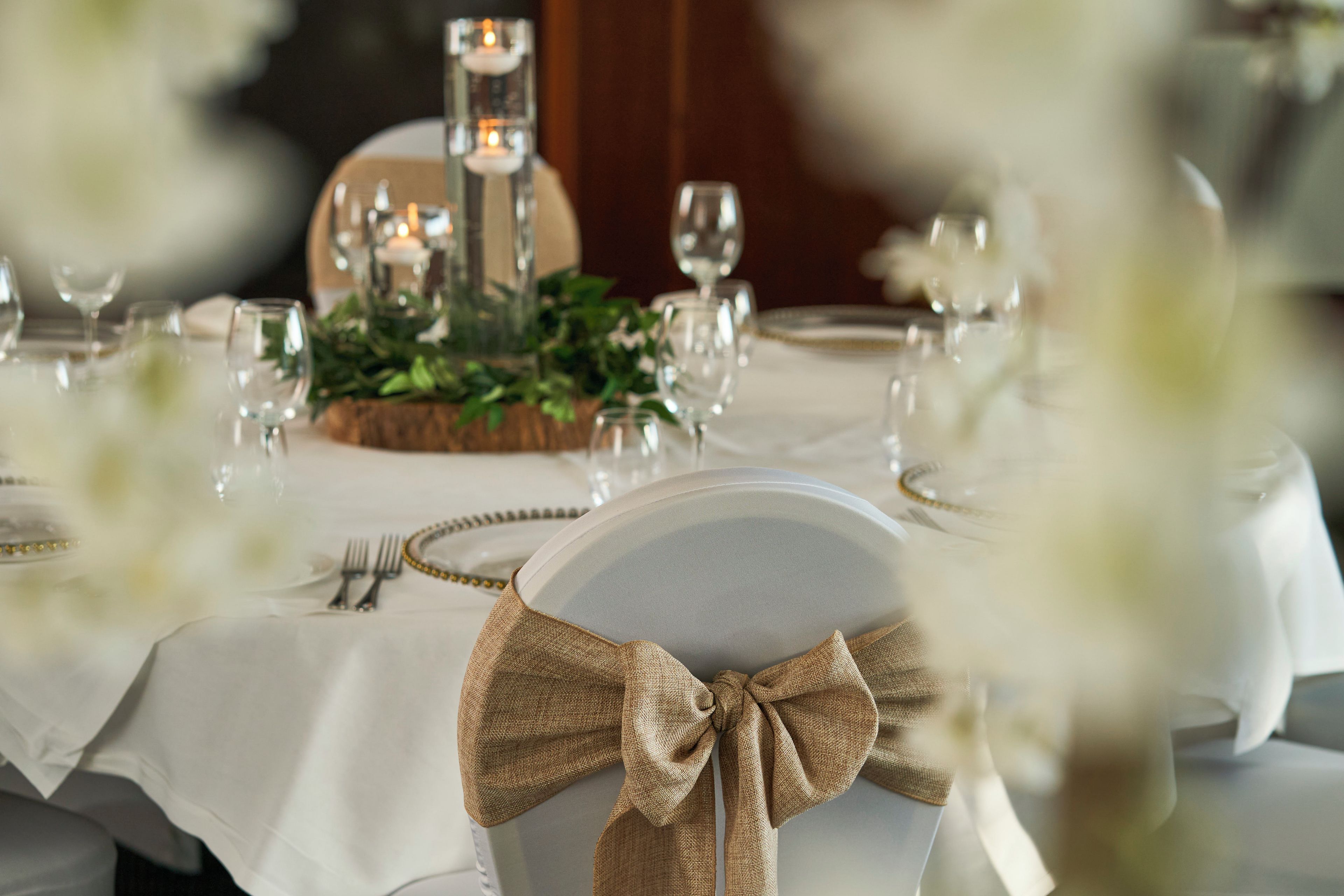 Una mesa vestida con manteles blancos, cubiertos y cristalería, con un centro de mesa decorativo y candelabros.