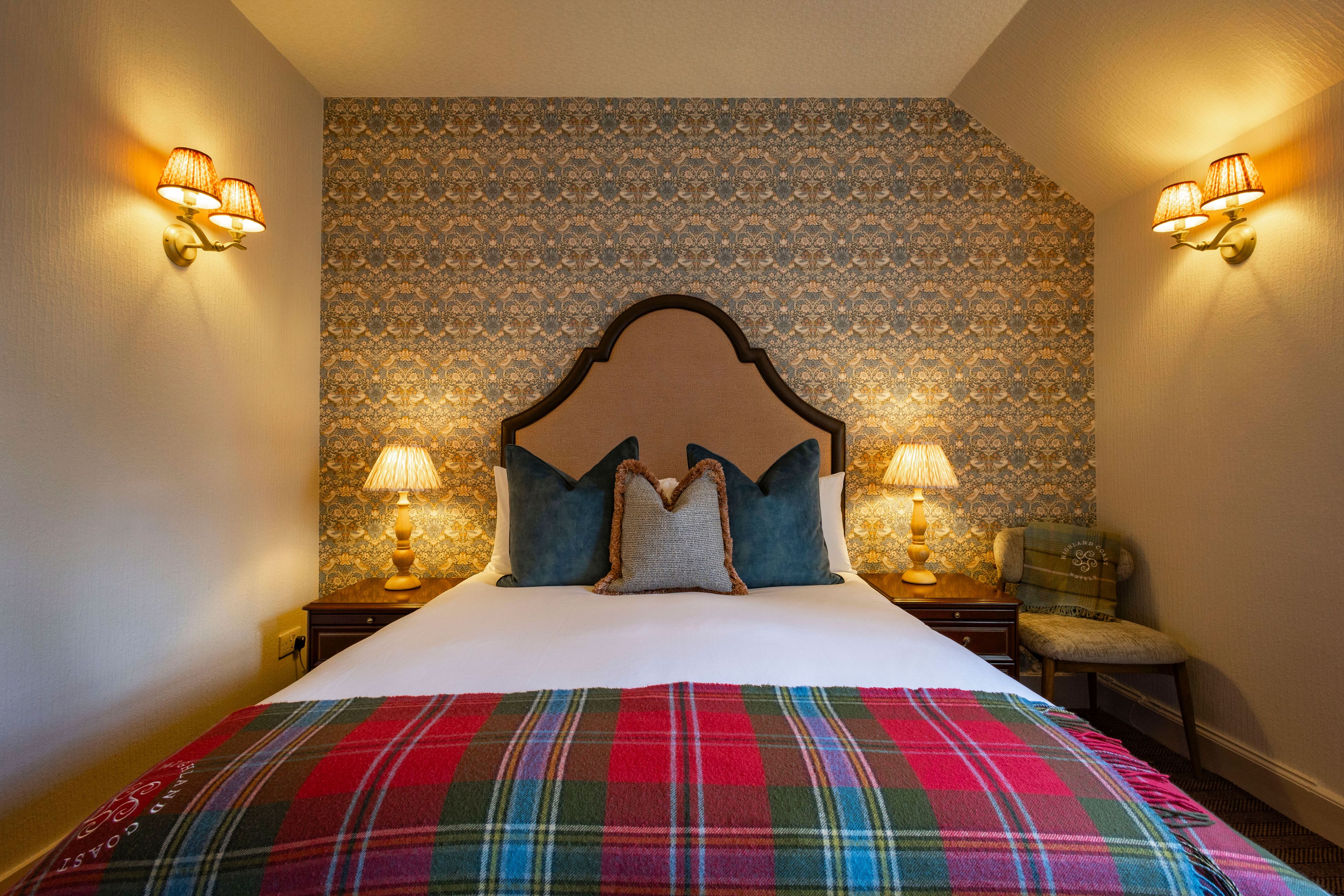 A hotel room with a large bed, two bedside lamps, a patterned wallpaper, and a plaid bedspread.