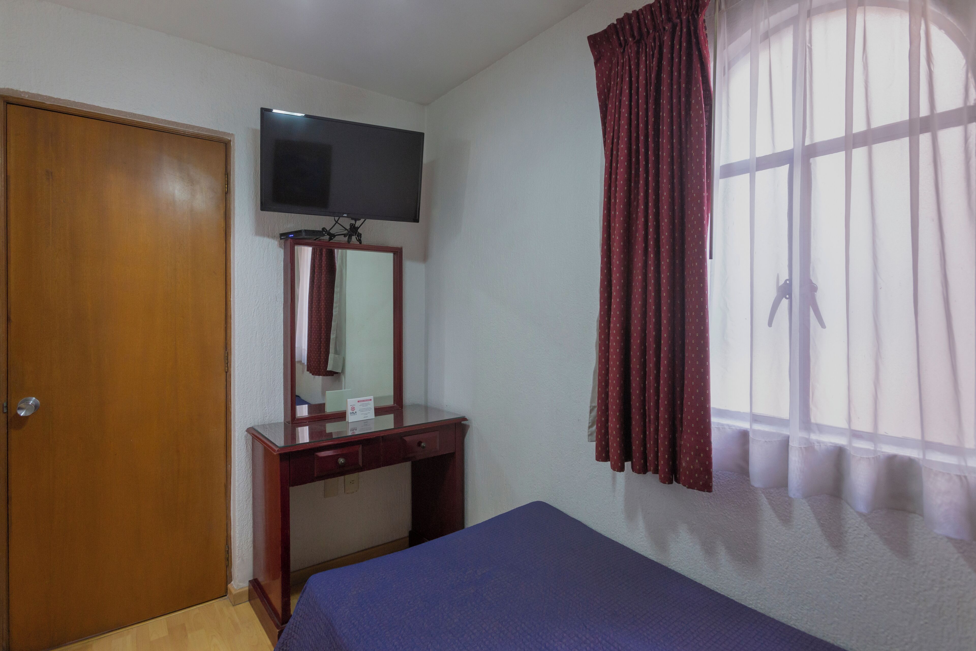 A room with a wooden wardrobe, a desk with a mirror, a bed, and a TV mounted on the wall.