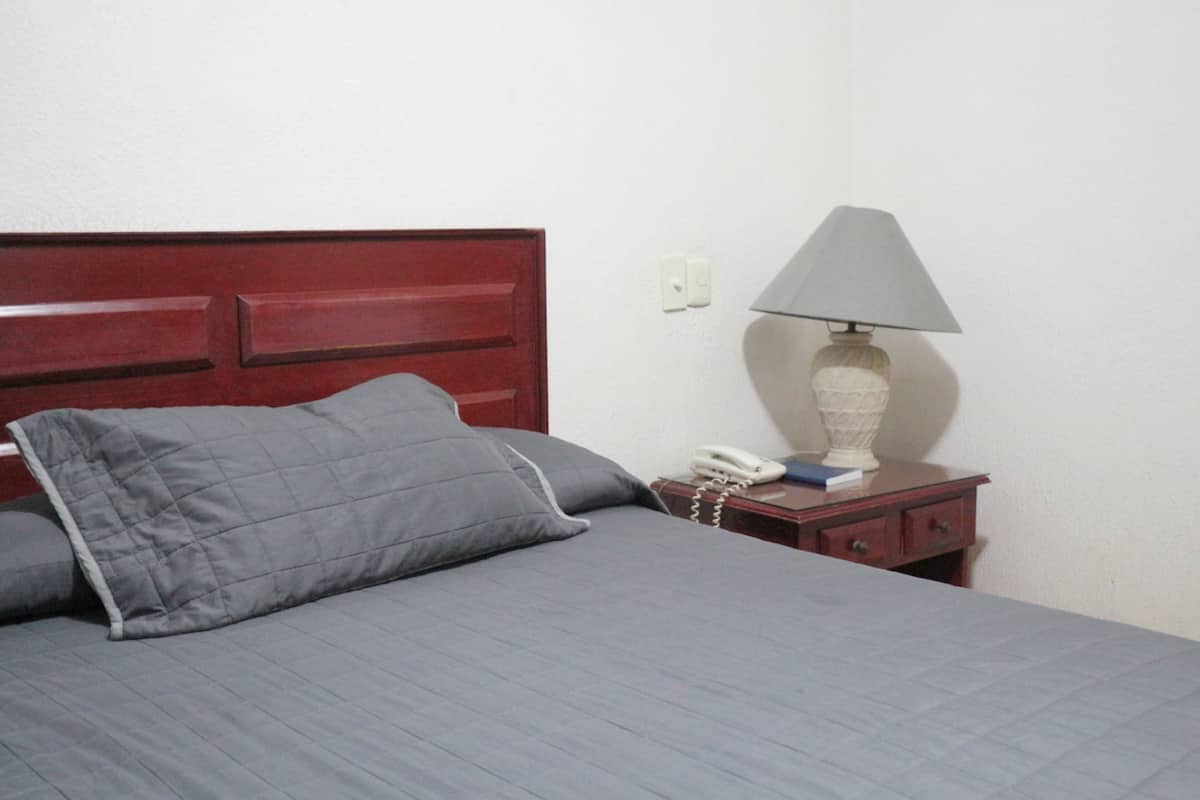 Una cama con colcha y almohadas grises, una mesita de noche con una lámpara, un teléfono y un libro.