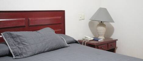 A bed with a grey quilt and pillows, a bedside table with a lamp, a phone, and a book.