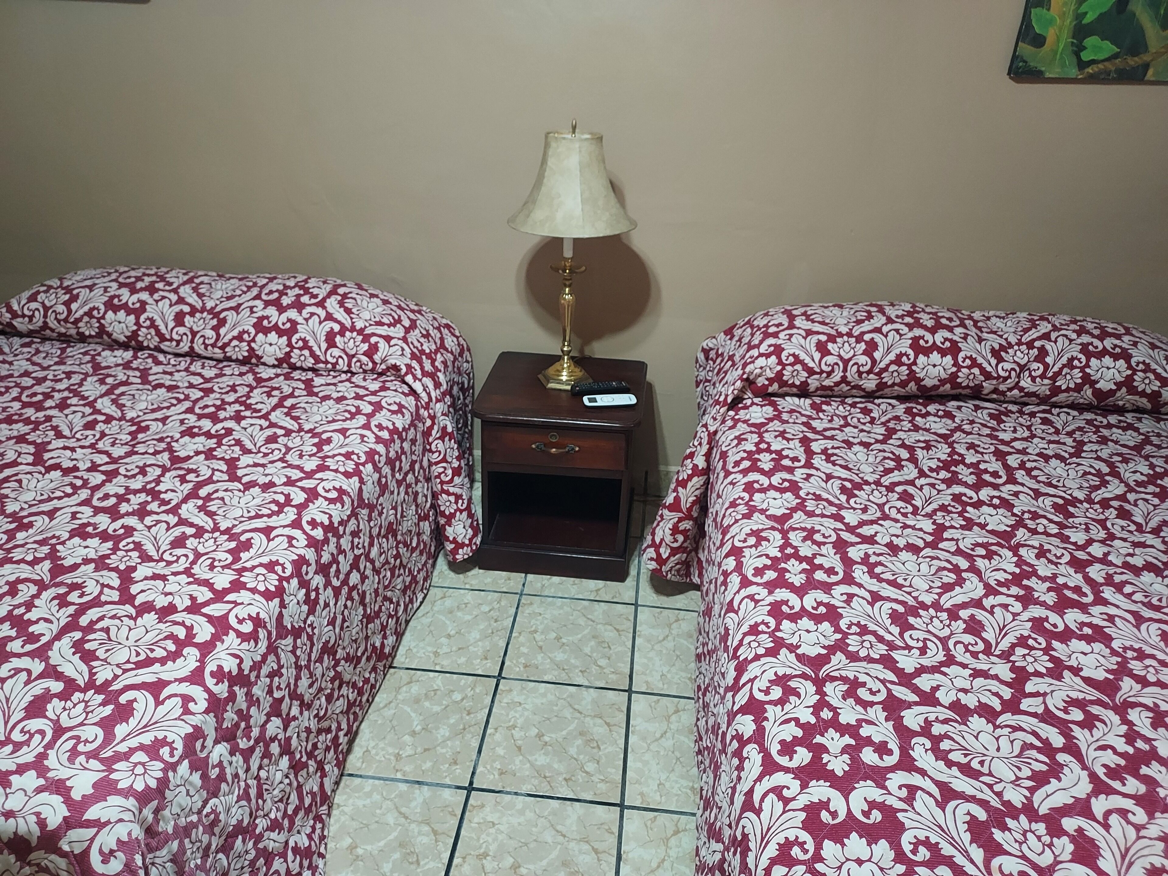 A room with two beds, each with a patterned bedspread, a nightstand with a lamp, and a small drawer.