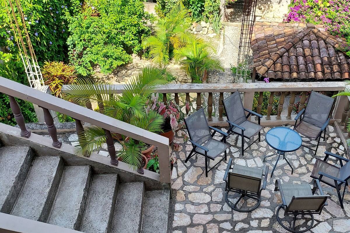A stone-floored outdoor patio with a round table and chairs, surrounded by lush greenery and a tiled roof structure.
