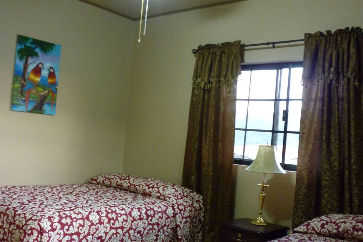 A room with two beds, a ceiling fan, a window with curtains, a painting on the wall, and a lamp on a bedside table.