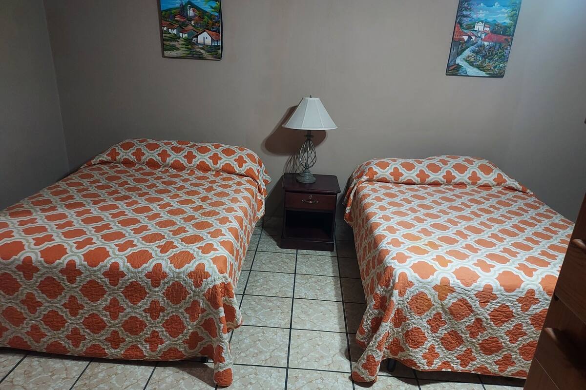 A room with two beds, a nightstand with a lamp, and two wall-mounted paintings.