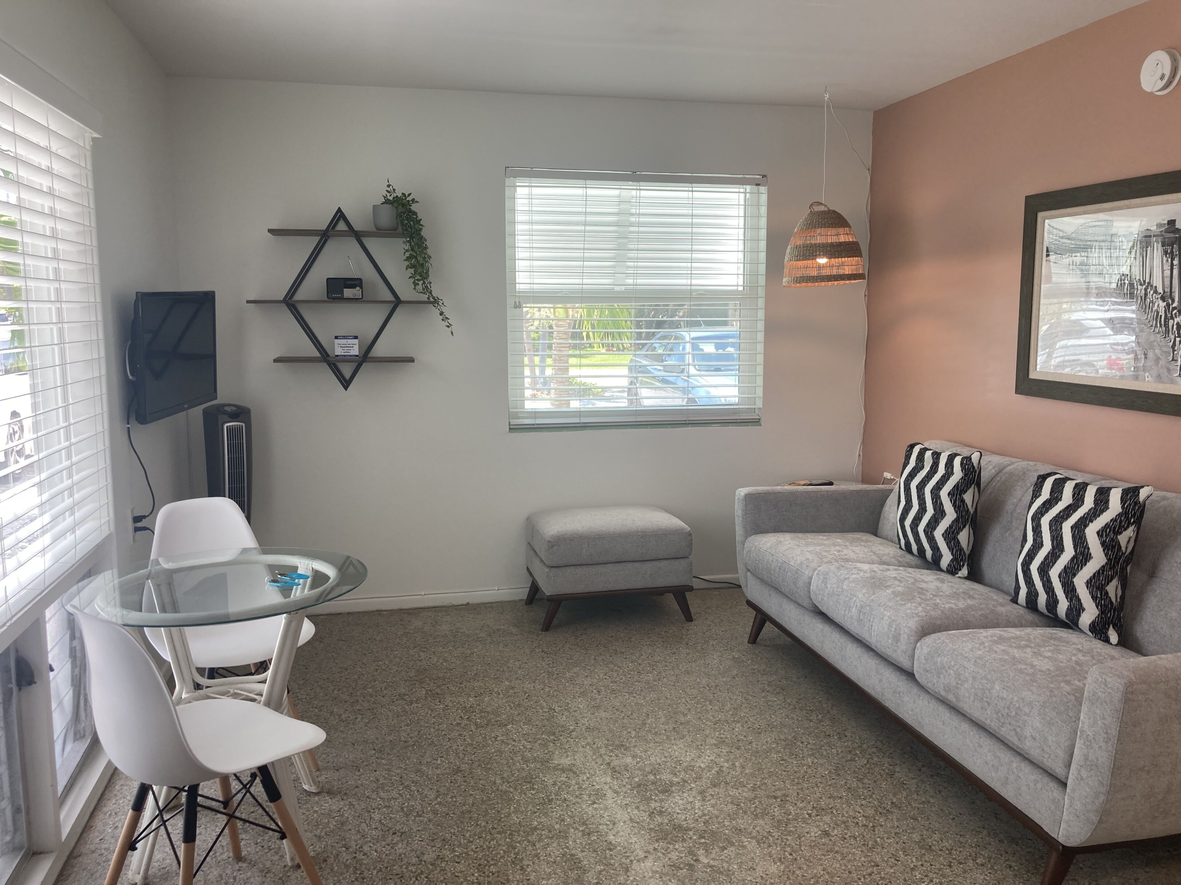 A living room with a grey sofa, white chairs, a glass table, a TV, a radiator, a window with blinds, a hanging lamp, and a framed picture on the wall.