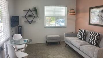 A living room with a grey sofa, white chairs, a glass table, a TV, a radiator, a window with blinds, a hanging lamp, and a framed picture on the wall.