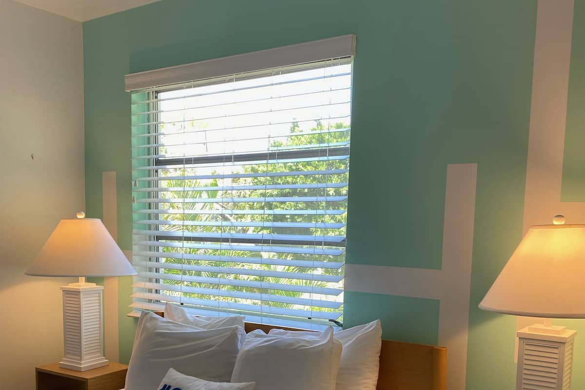 A neatly made bed with white bedding and pillows, including one with '#SSSL' text, flanked by two bedside tables with lamps, in a room with teal walls and a window with blinds.