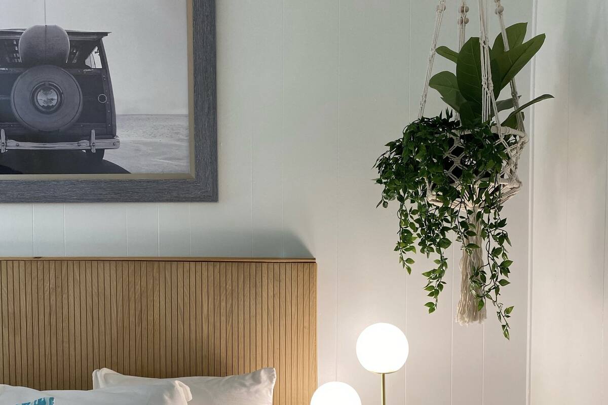 A neatly arranged bedroom with a wooden headboard, a bed with white and blue striped pillows, a bedside table with a lamp, and a hanging plant.