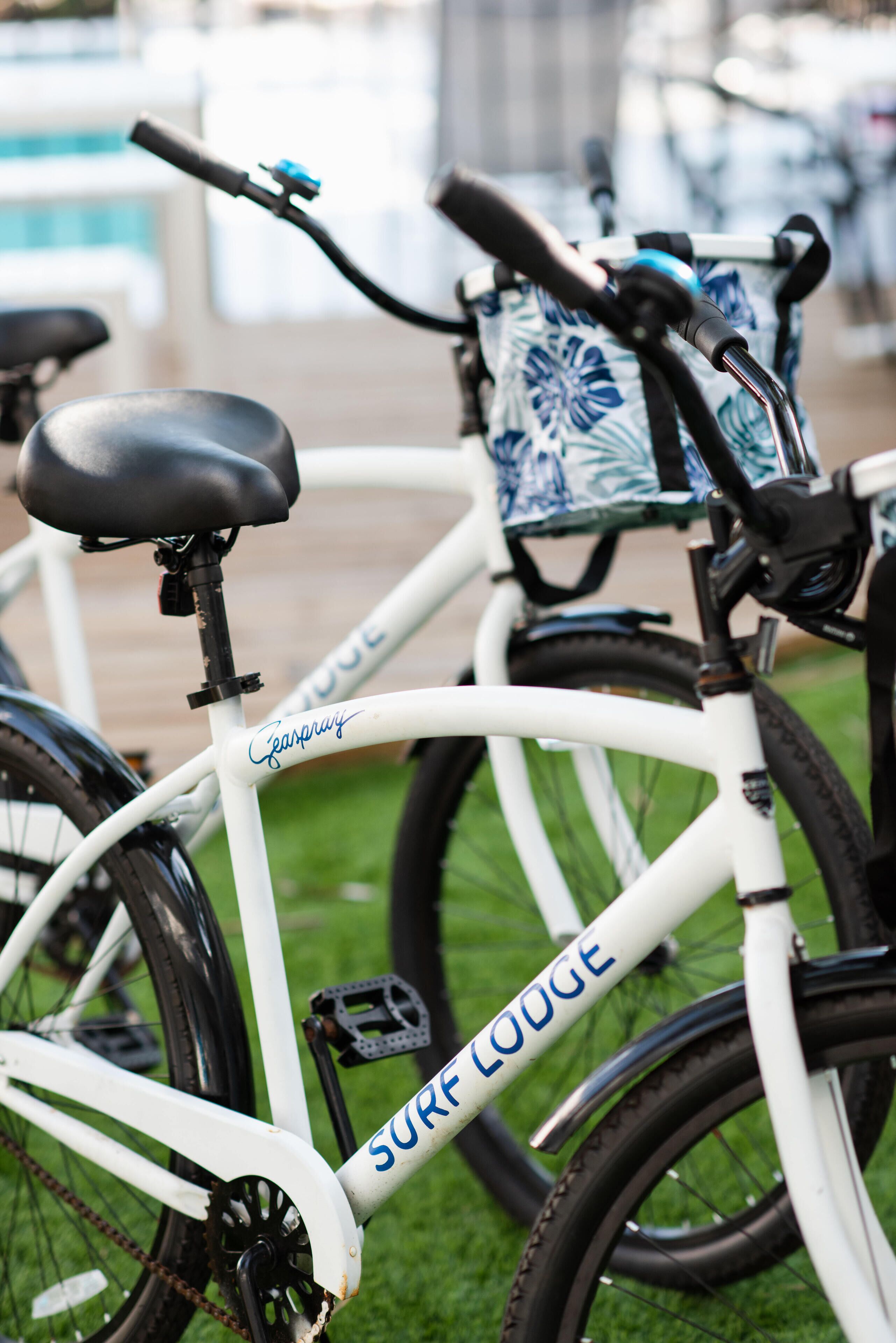 A white bicycle with "Surf Lodge" written on the frame, a black seat, and a blue and white patterned basket attached to the handlebars.