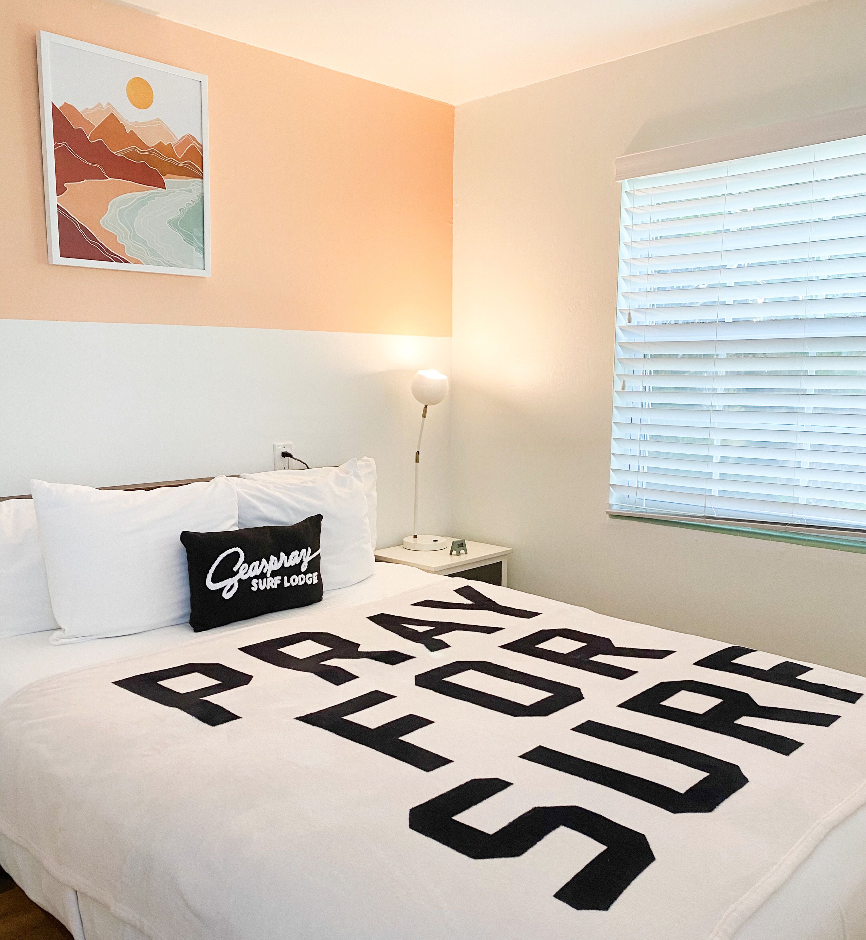 Un dormitorio con cama, almohadas, una mesita de noche, lámpara, ventana con persianas y una obra de arte en la pared.